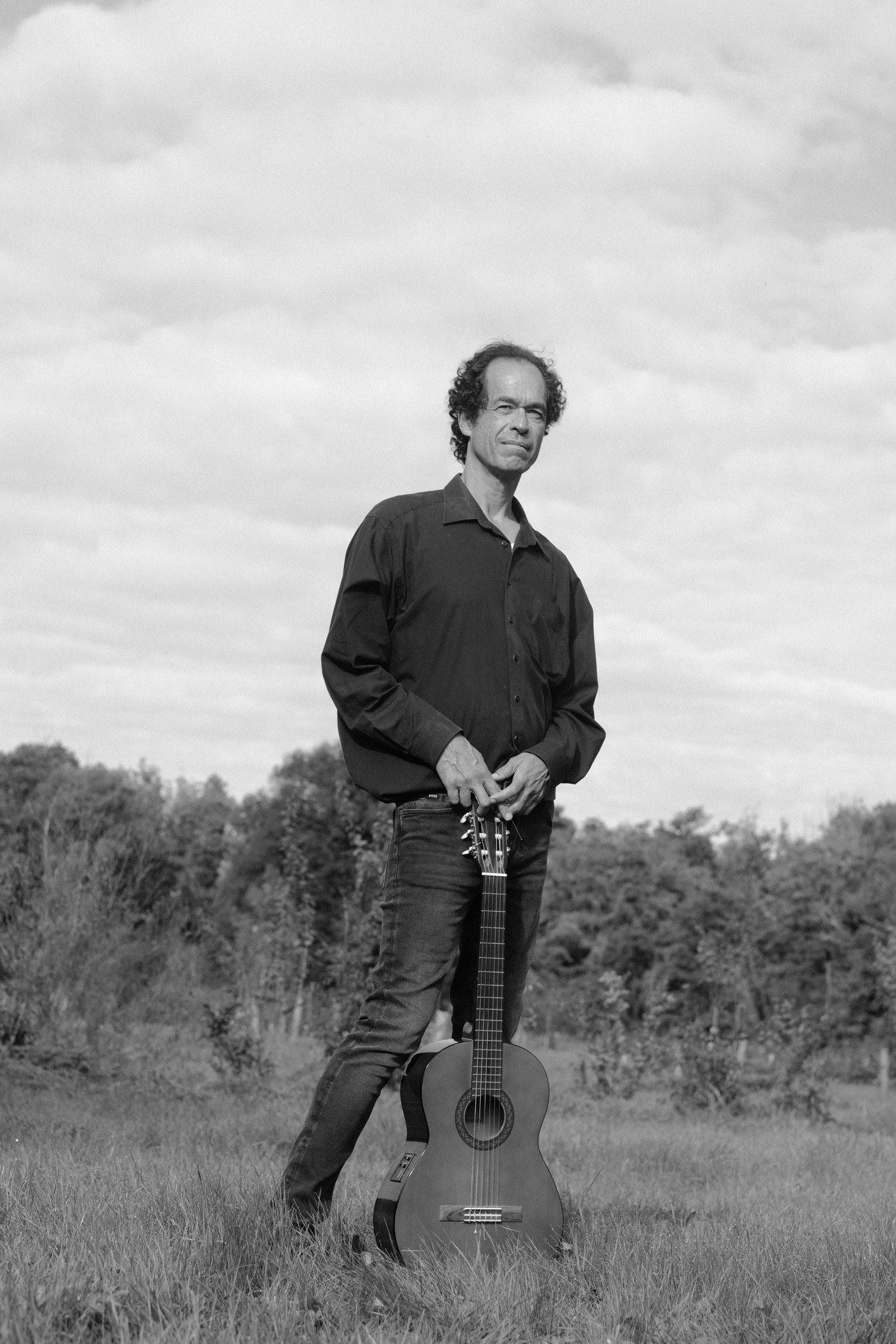 Black-and-white photo of Jérôme Inen standing in a meadow, leaning on a classical guitar